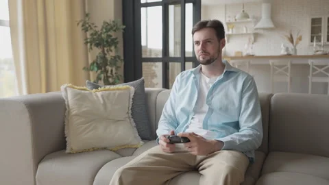 Focused Man Playing Video Game At Home Sitting On Couch With Serious Expression Stockbeeldmateriaal 311258814