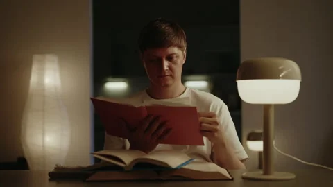 Focused man reading paper sitting at table book under soft glow of lamp in dimly Stock Footage 314269903