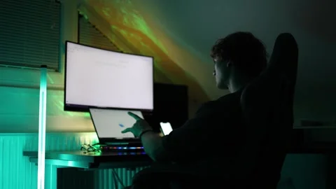 Focused man sits at desk using laptop and big screen in dark room illuminated by Stock Footage 316878200