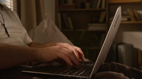 Focused Man Typing on Laptop Keyboard Working Remotely on Bed at Home Stock Footage 337500716