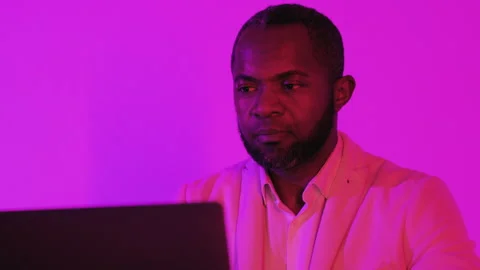 A focused man in a white suit jacket works on a laptop, illuminated by vibrant Stock Footage 295775554