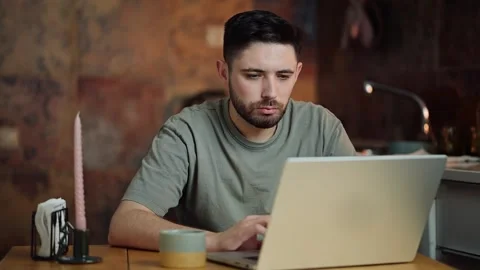 Focused man working on laptop at a cozy kitchen table with coffee and candle Stock Footage 300773631