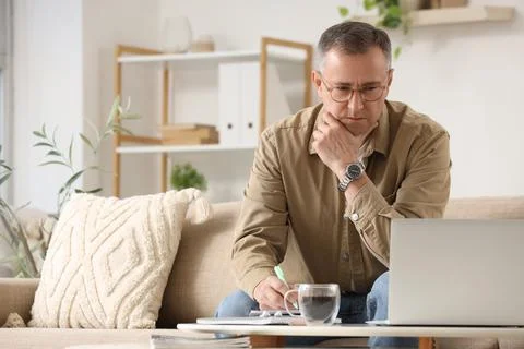 Focused man working remotely on laptop at home, taking notes and drinking cof Stock Photos