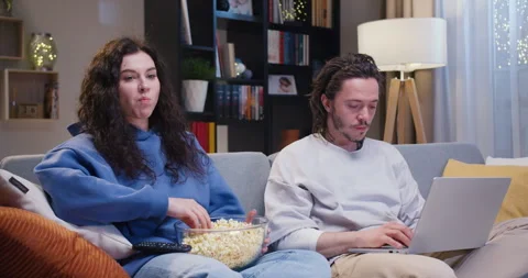 Focused man working remotely on modern laptop while woman eating popcorn and Stock Footage 244141784