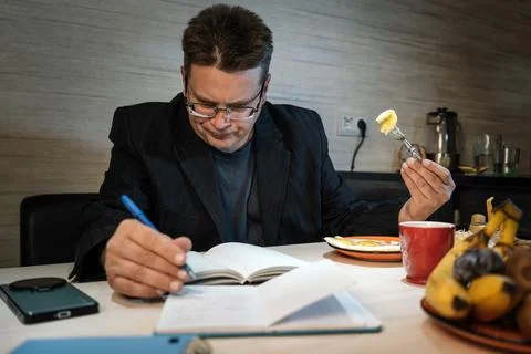 Focused man writing in a notebook with a pen while holding a fork with food d Stock Photos