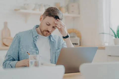 Focused man writing notes while sitting at kitchen table with laptop Stock Photos