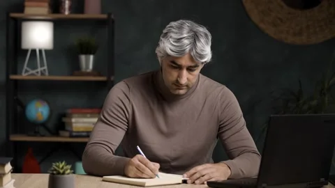 Focused Mature Grey Haired Man Writing in Notebook at Desk with Laptop Video stock 342236548