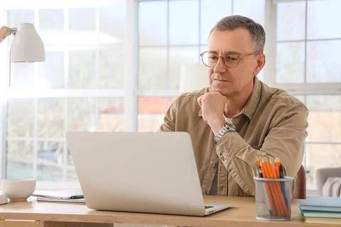 Focused mature man working remotely on laptop at home office desk, thoughtful Stock Photos