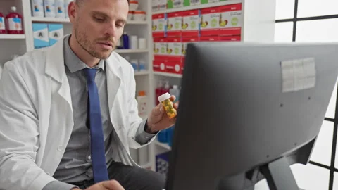 A focused pharmacist man examines medication while working on a computer in.. 動画素材 282580936