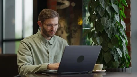 Focused Professional Working on Laptop in Cozy Cafe Setting Surrounded by Lush Stock Footage 344799770
