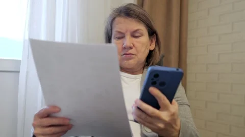 Focused senior woman comparing documents and smartphone data at home Stock Footage 306843777