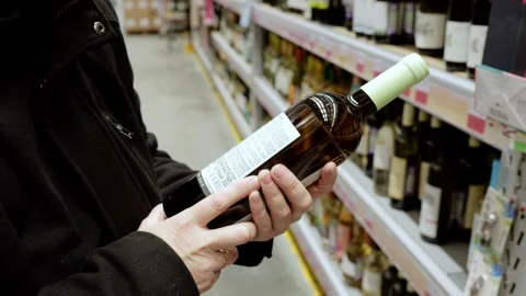 Focused shopper examining wine labels, standing in blurred supermarket aisle Stock Footage 328020385