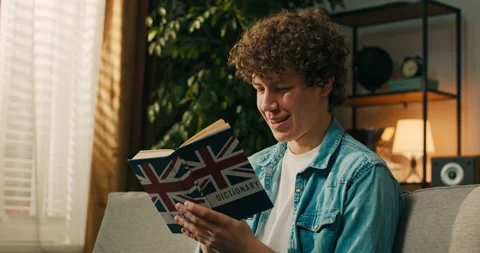 A focused, smiling student boy holds up an English dictionary and repeats new Stock Footage 218214917