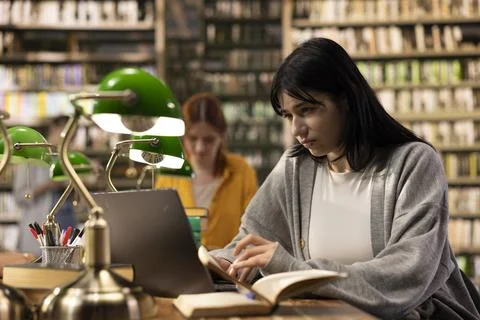 Focused student using laptop for note taking beside open book at library Stock Photos