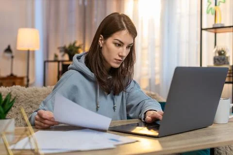 Focused woman freelancer reading documents and using laptop computer at table in Stock Photos