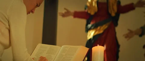 Focused woman reading of scripture beside burning candle and holy statue in Video stock 313698551
