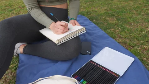 A focused woman sketching in a notebook while sitting cross-legged Stock Footage 264884599