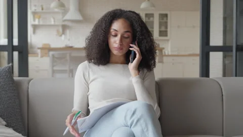 Focused Woman Taking Notes During Phone Call On Cozy Sofa In Bright Apartment Stockbeeldmateriaal 306699636