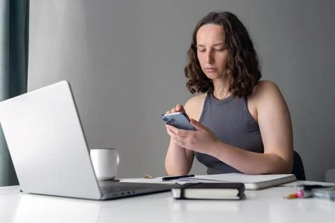 Focused woman using smartphone while working at home with laptop and notebook Stock Photos