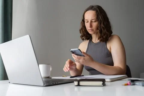Focused woman using smartphone while working at home with laptop and notebook Stock Photos