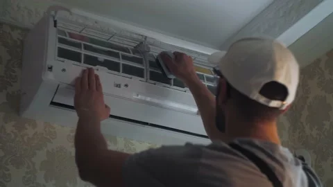 Focused young handyman cleans dust with brush on white air conditioner unit on Stock Footage 220834010