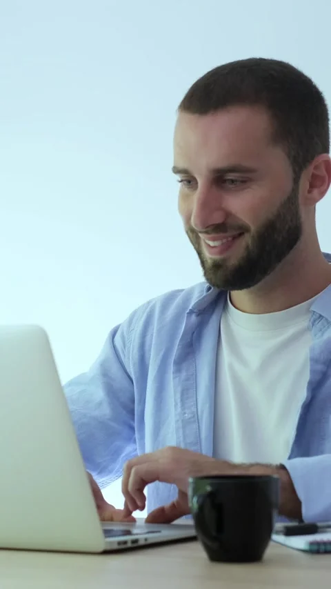 Focused young man with a beard sitting at a wooden desk, typing on a laptop Stock Footage 345114112