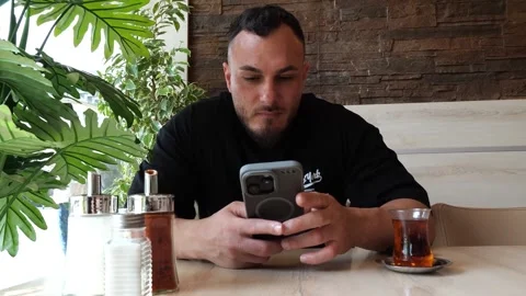 Focused young man sitting in a cafe using his smartphone and drinking tea 스톡 동영상 343574486