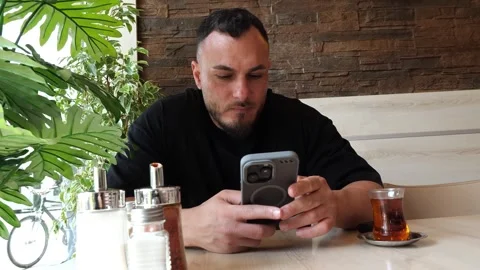 Focused young man sitting in a cafe using his smartphone and drinking tea Stock-Footage 343574527