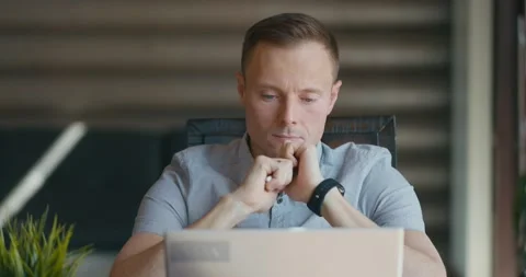 Focused young man is sitting in room at laptop. Holds pen in his hands. Looking Stock Footage 197816691