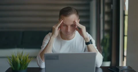 Focused young man is sitting at table in room with laptop. Rubs his temples with Stock Footage 197621401