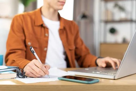 Focused young man sitting at table with laptop and making notes 스톡 사진