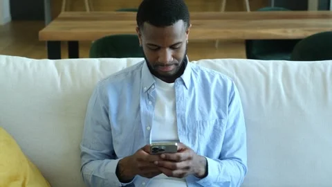 A focused young man uses his smartphone while relaxing on a comfortable sofa in Video stock 335242379