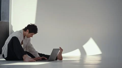 Focused young man using laptop on floor in bright minimal room with strong Stock Footage 345240921