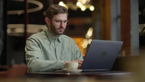 Focused Young Man Wearing Glasses Working on Laptop in Cozy Cafe Ambiance Stock Footage 344799726