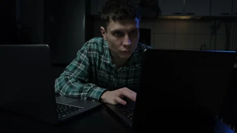 Focused young man working late at night on two laptops in a dimly lit room, inte Video stock 296959914