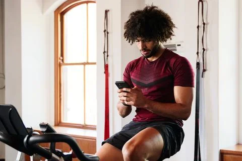 Focused young middle eastern man using mobile phone on exercise bike at the gym Stock Photos