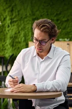 Focused young professional writing notes at desk with glasses, white shirt, warm Stock Photos