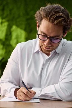 Focused young professional writing notes at desk with glasses, white shirt, warm Stock Photos