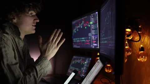 A focused young trader analyzing financial charts on multiple screens in a dark  Stock Footage 297112023