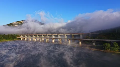 Fog covers a bridge over a river with mountains in the background during early Stock Footage 340929702