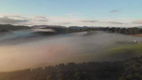 Foggy clouds wandering between hills during sunrise in New Zealand - aerial shot Video stock 153474572