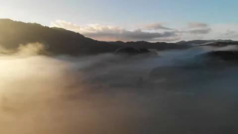 Foggy clouds wandering between hills during sunrise in New Zealand - aerial shot Video stock 153475587