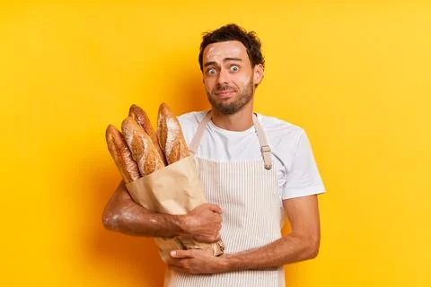 Food concept. Insecure handsome man baker in apron keeps bunch of fresh baked Stock Photos