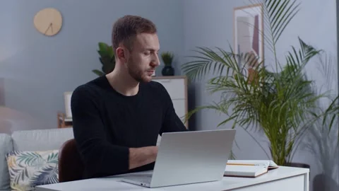 Footage of young attractive man with beard sitting at desk with laptop, takes Stock Footage 189028015