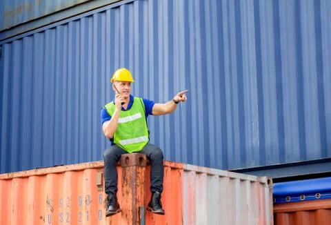 Foreman worker in hardhat sitting on container box talks on two-way radio Fotos de archivo