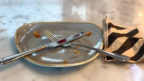 Fork and knife cross on gray plate, crumbs scattered, napkin rests beside after Stock Photos