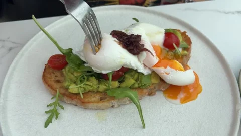 Fork breaking poached eggs with runny yolk on avocado toast Stock-Footage 347416192