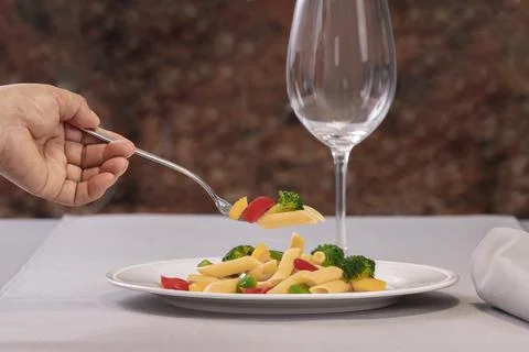 A fork with pasta and vegetables held by a hand over a plate of pasta salad Stock Photos