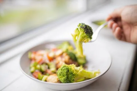 Fork picking broccoli from a broccoli and chicken salad next to a window Stock Photos