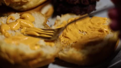 Fork spreading cheese, mustard, and yellow sauce on white burger bun Stock Footage 310902965